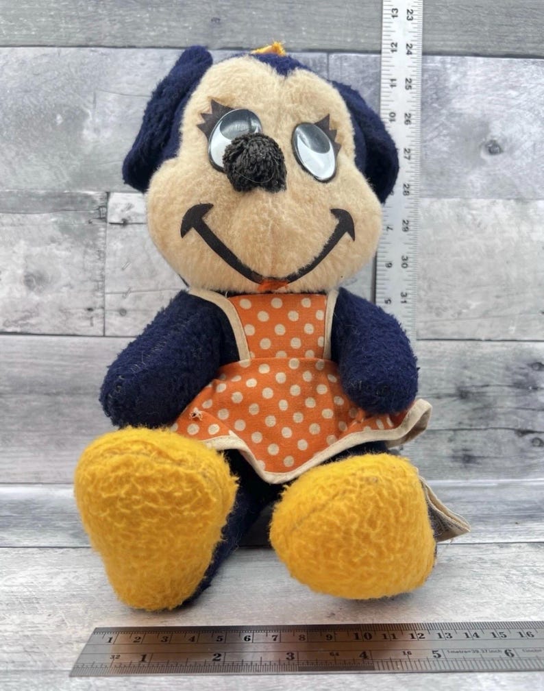 May include: Vintage plush dog toy with a cream-colored face, black nose, and dark blue ears and limbs. It wears an orange polka-dot apron and has yellow feet. The toy is approximately 10 inches tall.