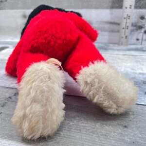 May include: A plush Santa Claus doll, viewed from the feet up. The doll's body is red with a black hat. The feet are white and fluffy, with a cream-coloured tag attached to one foot. The doll is lying on a grey surface.