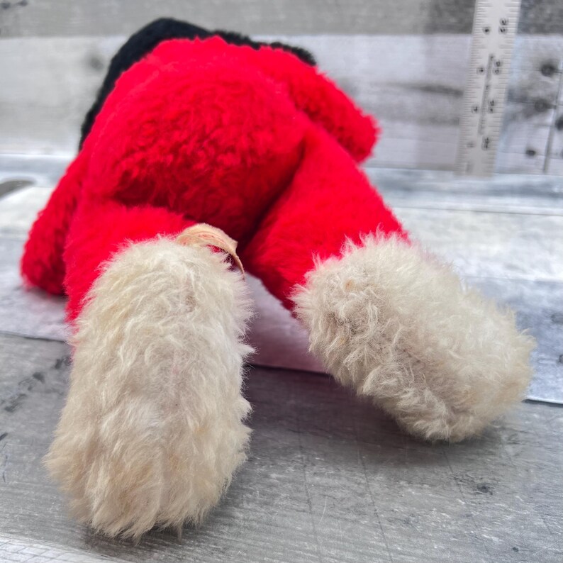 May include: A plush Santa Claus doll, viewed from the feet up. The doll's body is red with a black hat. The feet are white and fluffy, with a cream-coloured tag attached to one foot. The doll is lying on a grey surface.
