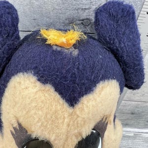 May include: Close-up of a vintage plush toy with a dark blue head and ears, and a tan face. The toy has large, black and white eyes and a small tuft of yellow fabric on top of its head. The toy appears to be well-loved.