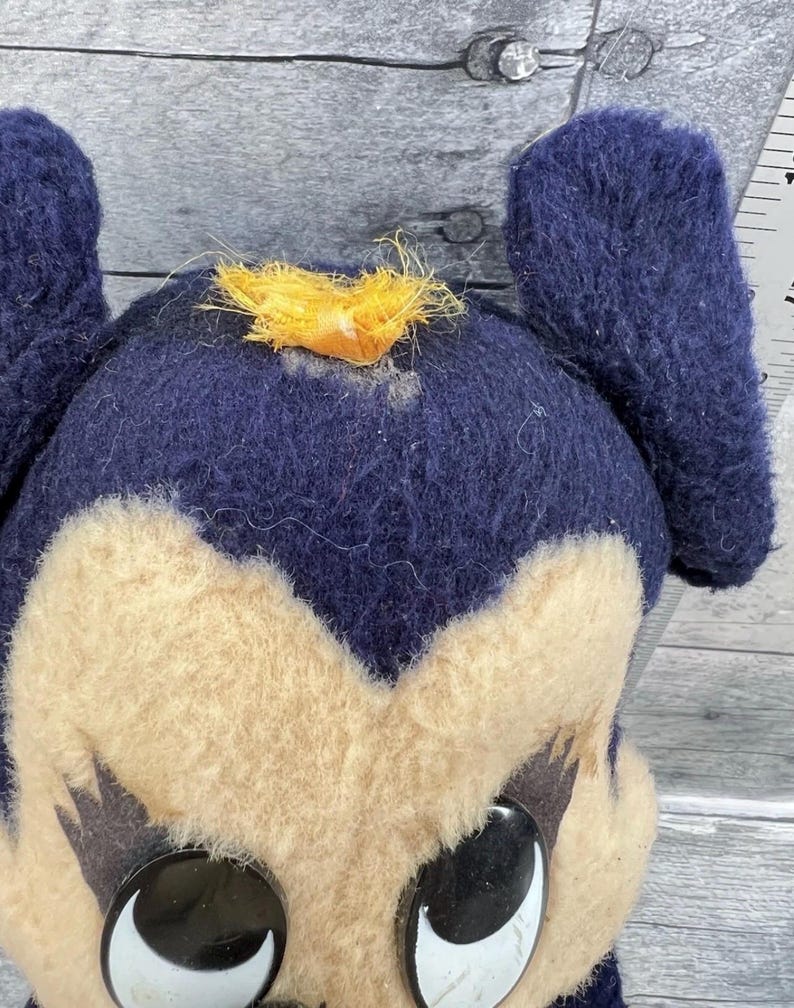 May include: Close-up of a vintage plush toy with a dark blue head and ears, and a tan face. The toy has large, black and white eyes and a small tuft of yellow fabric on top of its head. The toy appears to be well-loved.