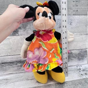 Puede incluir: Mu&ntilde;eca de peluche de Minnie Mouse con un vestido con estampado floral, zapatos amarillos y un lazo. La mu&ntilde;eca mide aproximadamente 30 cm de alto. Las orejas de la mu&ntilde;eca son negras y la cara es de color canela con ojos y nariz negros.