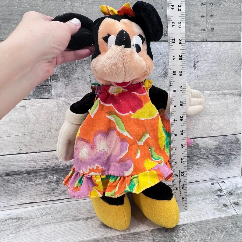Puede incluir: Mu&ntilde;eca de peluche de Minnie Mouse con un vestido con estampado floral, zapatos amarillos y un lazo. La mu&ntilde;eca mide aproximadamente 30 cm de alto. Las orejas de la mu&ntilde;eca son negras y la cara es de color canela con ojos y nariz negros.
