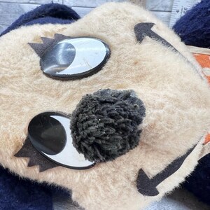 May include: Close-up of a vintage plush toy face with large, reflective eyes and a fluffy, dark pom-pom nose. The toy has a cream-colored face with black and white accents and a navy blue and orange patterned border.