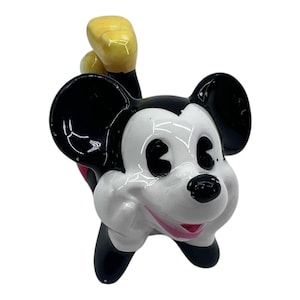May include: A ceramic figurine of Mickey Mouse in a playful pose. The figure features black ears, a white face, and a red shorts. The hands are yellow, and the figure has a glossy finish. A classic Disney collectible.