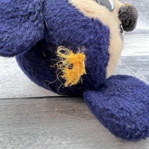 May include: A close-up of a vintage stuffed owl toy. The owl has a navy blue body, a beige face, and black and white eyes. The owl's wing is visible, and there is a patch of orange thread on the body.