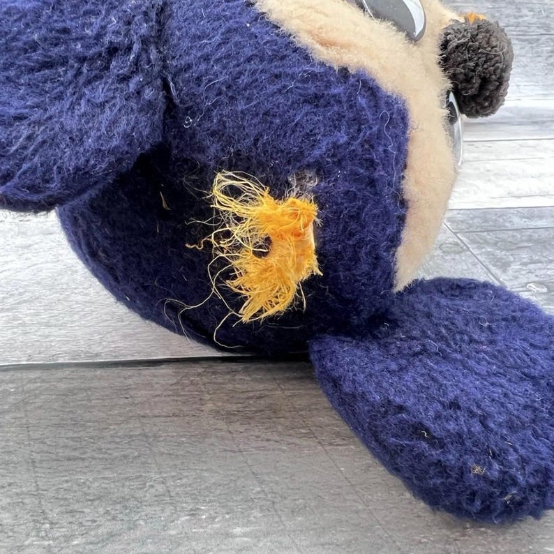 May include: A close-up of a vintage stuffed owl toy. The owl has a navy blue body, a beige face, and black and white eyes. The owl's wing is visible, and there is a patch of orange thread on the body.