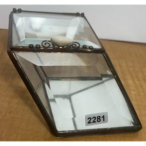 May include: A clear glass box with a mirrored interior and a decorative metal trim. The box has a small white stone on the top and a sticker with the number 2281 on the bottom.