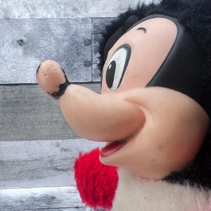 May include: A vintage plush toy depicting a cartoon character's face. The character has a large, pink nose, a red mouth, and a black head with fluffy black hair. The toy's design is reminiscent of classic cartoon aesthetics.