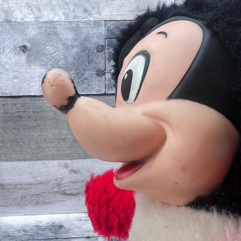 May include: A vintage plush toy depicting a cartoon character's face. The character has a large, pink nose, a red mouth, and a black head with fluffy black hair. The toy's design is reminiscent of classic cartoon aesthetics.