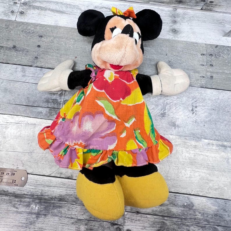 Puede incluir: Una mu&ntilde;eca de peluche de Minnie Mouse con orejas negras, una cara bronceada y un vestido floral colorido. La mu&ntilde;eca tiene guantes blancos, zapatos amarillos y un peque&ntilde;o lazo en la cabeza. El vestido presenta flores naranjas, rosas y verdes.