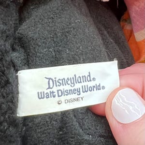 Puede incluir: Una etiqueta rectangular blanca con las palabras "Disneyland" y "Walt Disney World" impresas en escritura azul oscuro. La etiqueta tambi&eacute;n incluye el logotipo de Disney. La etiqueta est&aacute; cosida a una tela gris oscuro texturizada.