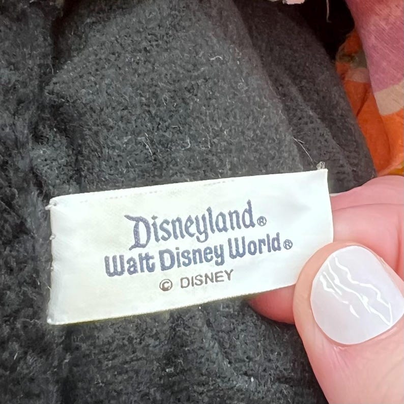 Puede incluir: Una etiqueta rectangular blanca con las palabras "Disneyland" y "Walt Disney World" impresas en escritura azul oscuro. La etiqueta tambi&eacute;n incluye el logotipo de Disney. La etiqueta est&aacute; cosida a una tela gris oscuro texturizada.