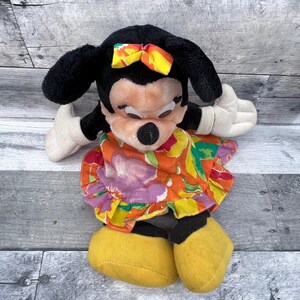 Puede incluir: Un peluche de Minnie Mouse con orejas negras, nariz negra y guantes blancos. Lleva un vestido floral colorido y un lazo amarillo y naranja. El peluche tiene zapatos amarillos.