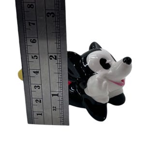 May include: A black, white, and pink ceramic figurine of a cartoon dog, approximately 2 inches tall. The figurine has a glossy finish and is positioned next to a ruler for scale. The dog has large ears and a smiling expression.