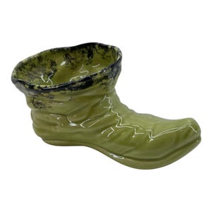 May include: A ceramic, green boot-shaped planter with a glossy finish. The top rim is black with a speckled pattern. The boot has a pointed toe and a curved shape, suitable for small plants or decorative use.