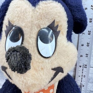 May include: A vintage plush toy featuring a cartoon dog face with large, reflective eyes and a black pom-pom nose. The dog has a cream-colored face, dark blue ears, and an orange polka-dot bib. The toy has a yellow tuft of hair.