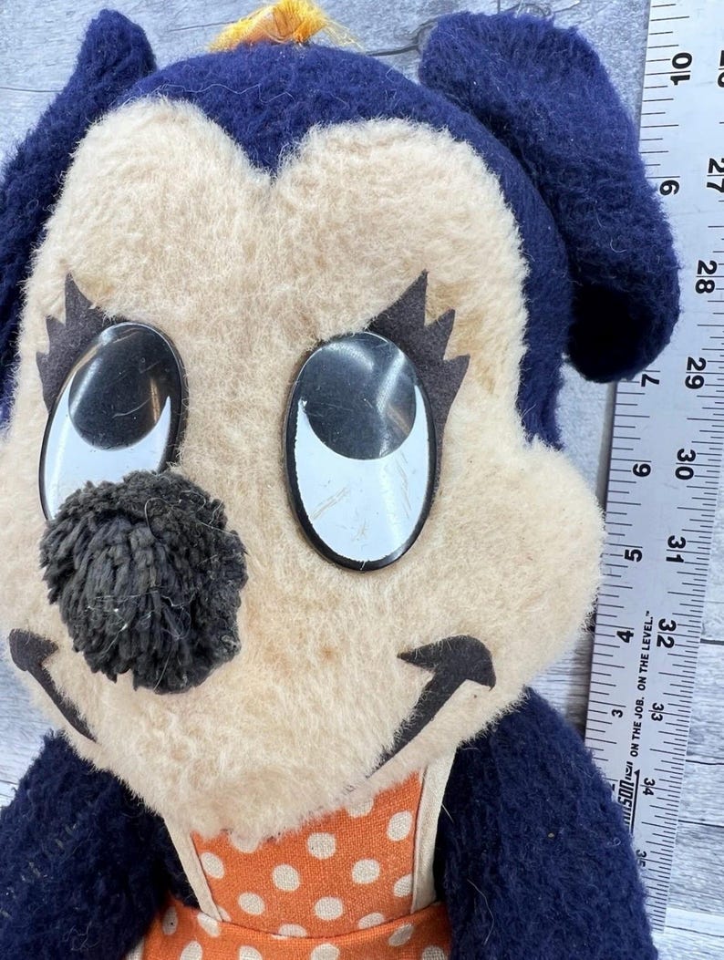 May include: A vintage plush toy featuring a cartoon dog face with large, reflective eyes and a black pom-pom nose. The dog has a cream-colored face, dark blue ears, and an orange polka-dot bib. The toy has a yellow tuft of hair.