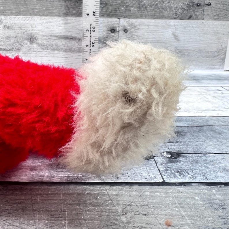 May include: Close-up of a plush toy's foot and leg. The leg is a vibrant red, while the foot is a fluffy, off-white colour. The toy's fur appears soft and textured. The background is a light grey wooden surface.