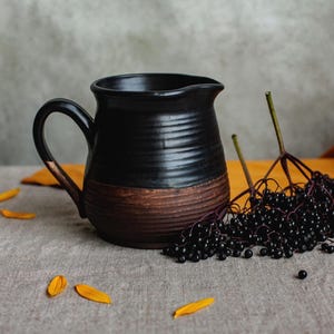 May include: A black ceramic jug with a brown base and handle. The jug is next to a cluster of dark berries and yellow flower petals on a neutral linen surface. The jug has a rustic, textured appearance.