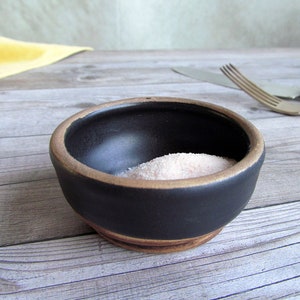 May include: A small black ceramic bowl with a brown rim, filled with pink Himalayan salt. The bowl is sitting on a wooden surface.