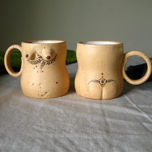 May include: Two beige ceramic mugs with handles, each featuring a unique design. One mug has a breast design with a leaf pattern, while the other has a stylized bottom design. The mugs are on a light grey surface.