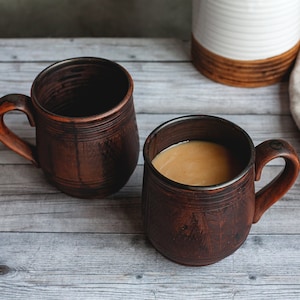 May include: Two rustic brown ceramic mugs with handles, one filled with a light brown liquid, likely coffee or tea. The mugs sit on a weathered, light gray wooden surface, with a neutral-toned cloth and a white and brown ceramic container in the background.