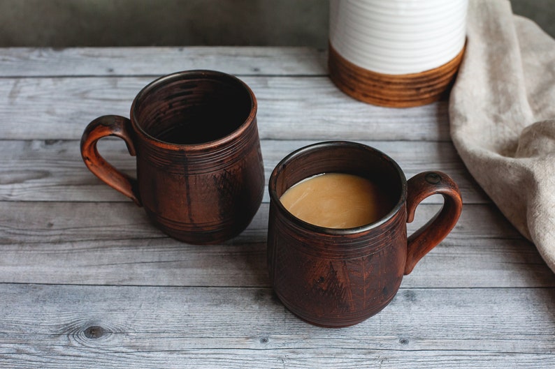 May include: Two rustic brown ceramic mugs with handles, one filled with a light brown liquid, likely coffee or tea. The mugs sit on a weathered, light gray wooden surface, with a neutral-toned cloth and a white and brown ceramic container in the background.
