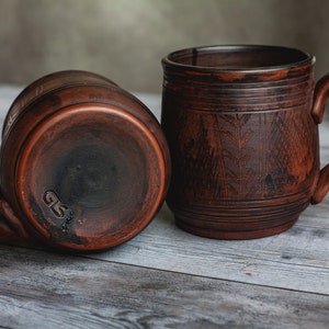 May include: Two rustic, brown ceramic mugs with handles. One mug is upright, the other is on its side. The mugs have a textured surface and a carved leaf design. The bottom of one mug has the initials "GS" stamped on it.