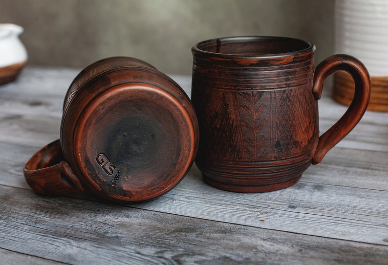 May include: Two rustic, brown ceramic mugs with handles. One mug is upright, the other is on its side. The mugs have a textured surface and a carved leaf design. The bottom of one mug has the initials "GS" stamped on it.
