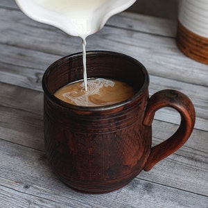 May include: A brown ceramic mug being filled with cream from a white pitcher. The mug has a large handle and a textured surface. The coffee and cream swirl together, creating a light brown color.
