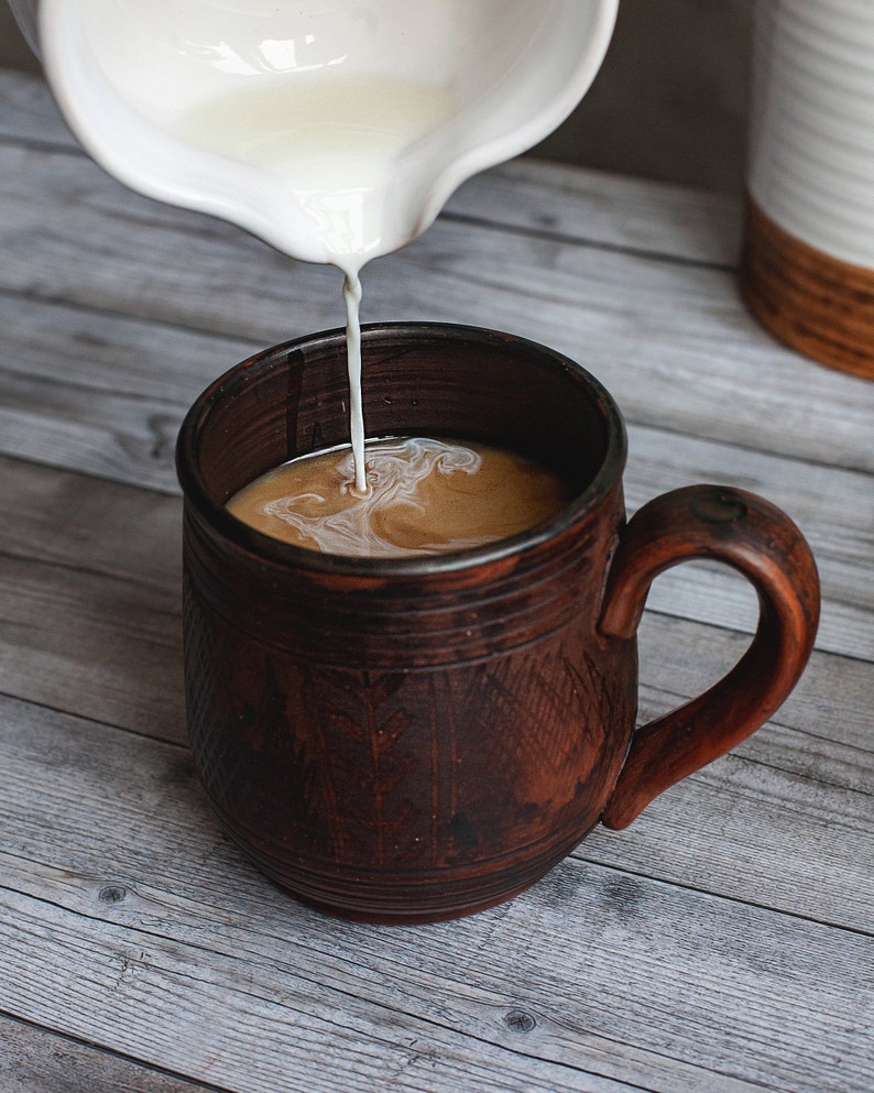 May include: A brown ceramic mug being filled with cream from a white pitcher. The mug has a large handle and a textured surface. The coffee and cream swirl together, creating a light brown color.