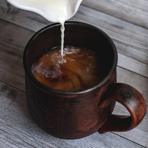 May include: A brown ceramic mug with a textured surface is filled with coffee as cream is poured from a white creamer. The mug sits on a gray wooden surface. The cream swirls into the coffee.