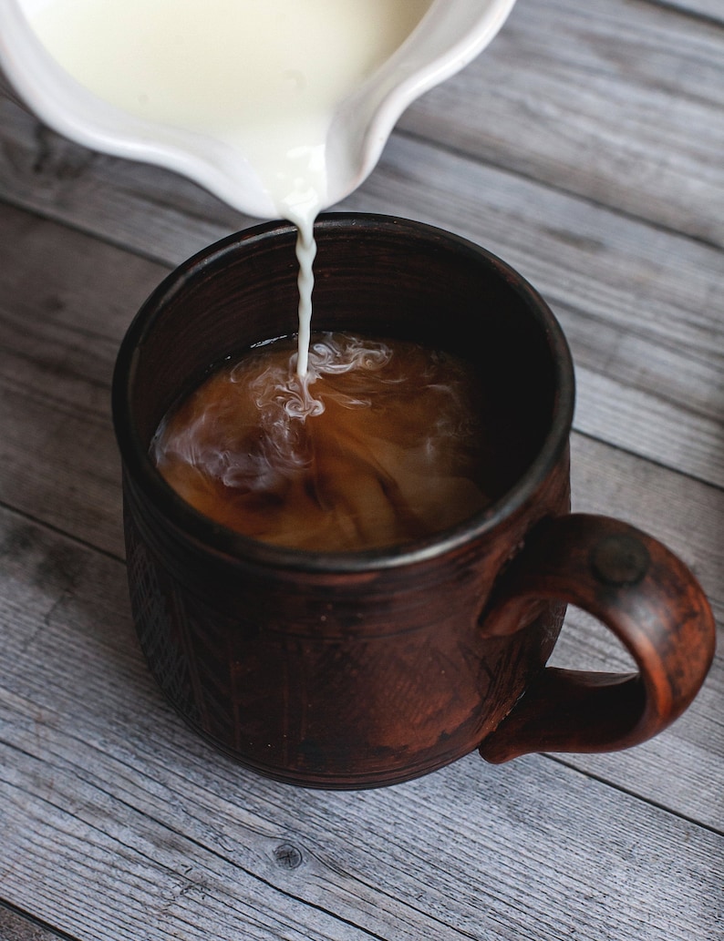May include: A brown ceramic mug with a textured surface is filled with coffee as cream is poured from a white creamer. The mug sits on a gray wooden surface. The cream swirls into the coffee.