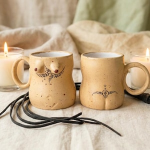 Könnte beinhalten: Zwei beige Keramikbecher mit gesprenkelter Oberfläche, jeder mit einer einzigartigen Körperform und einem Henkel. Ein Becher zeigt ein Brustdesign mit einem Blattmuster, der andere ein stilisiertes Design des unteren Rückens. Kerzen und eine schwarze Lederschnur sind ebenfalls vorhanden.