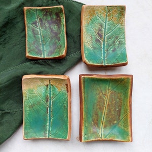 Puede incluir: Cuatro pequeños platos de cerámica con un diseño de hoja verde y marrón. Los platos son rectangulares y tienen un borde ligeramente curvado.