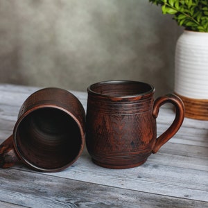 May include: Two rustic brown ceramic mugs with handles, one upright and one on its side, sit on a weathered wooden surface. The mugs have a textured surface and decorative etched details. A white vase with greenery is in the background.