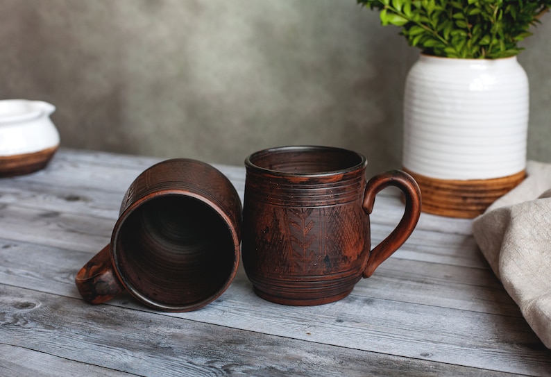 May include: Two rustic brown ceramic mugs with handles, one upright and one on its side, sit on a weathered wooden surface. The mugs have a textured surface and decorative etched details. A white vase with greenery is in the background.