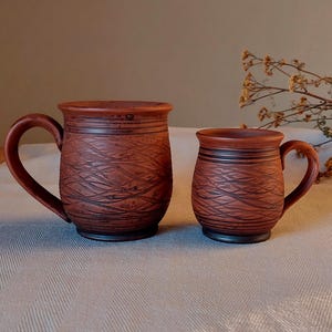May include: Two handmade, rustic-style ceramic mugs. The mugs are brown with a textured, carved design. The larger mug has a curved handle, and the smaller mug has a similar handle. The mugs are on a light-colored tablecloth.