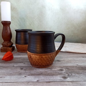 May include: Two handmade ceramic mugs with a black upper half and a textured, brown lower half. One mug has a handle. A wooden candlestick and a dried orange flower are also visible.