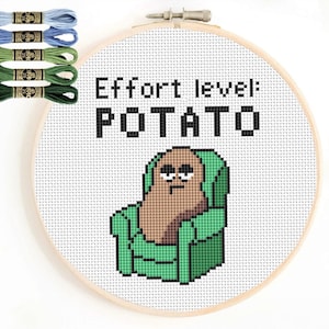 May include: A cross-stitch hoop featuring a pixelated potato character in a green armchair, with the text "Effort level: POTATO". The image includes embroidery floss in shades of blue and green.