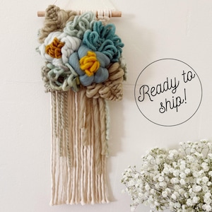 May include: A wall hanging featuring a wooden dowel with cream, sage green, teal, and mustard yellow yarn flowers and textured fibre elements. Long cream-coloured yarn strands hang from the bottom. The text Ready to ship! is visible.
