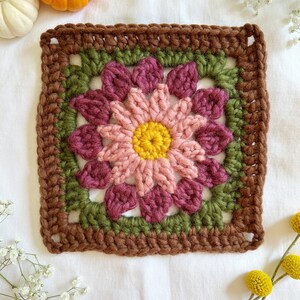 May include: A crocheted square with a pink and burgundy flower in the center. The flower has a yellow center and is surrounded by green leaves. The square is bordered with brown yarn.