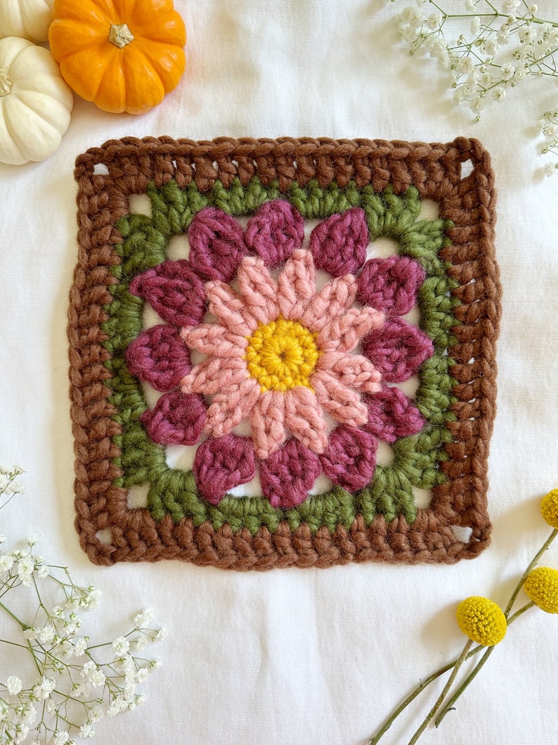 May include: A crocheted square with a pink and burgundy flower in the center. The flower has a yellow center and is surrounded by green leaves. The square is bordered with brown yarn.
