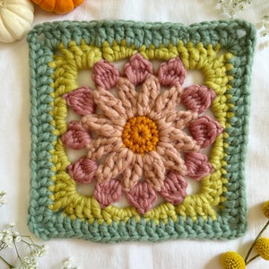 May include: A crocheted square with a flower design in shades of pink, yellow, and green. The flower has a center of orange yarn.