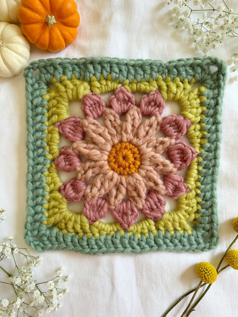 May include: A crocheted square with a flower design in shades of pink, yellow, and green. The flower has a center of orange yarn.