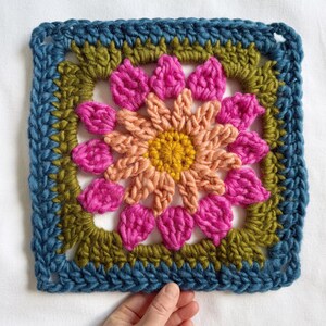 May include: A crocheted square with a colorful flower design. The flower has a yellow center, peach petals, and pink outer petals. The square is bordered with teal, green, and brown yarn.