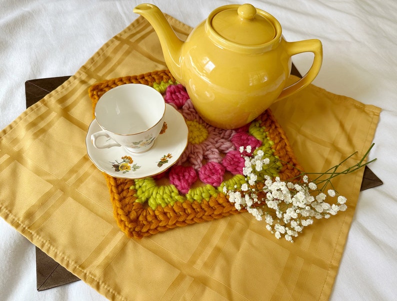 May include: A yellow teapot and a white teacup with floral designs are placed on a crocheted coaster with pink, green, and orange accents. The coaster is on a yellow tablecloth with a grid pattern. A sprig of white baby's breath flowers is in the foreground.