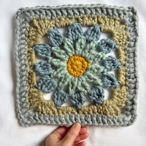May include: A crocheted square with a flower design in shades of blue, green, and yellow. The flower has a yellow center and is surrounded by blue and green petals.