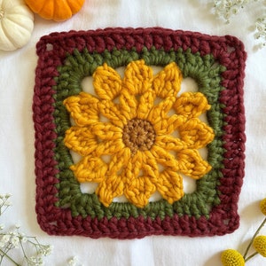 May include: A crocheted sunflower square with a burgundy border, a green inner border, and a yellow sunflower in the center.